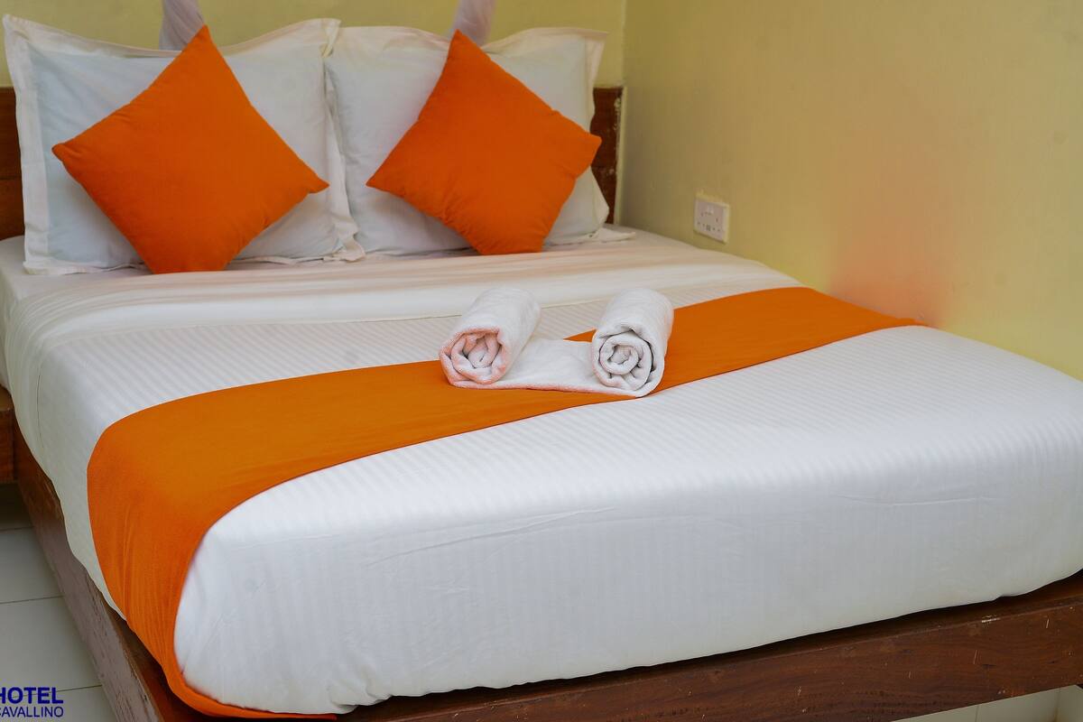 Cama bem arrumada com roupa de cama branca e laranja, dois toalhas enroladas e cabeceira de madeira.