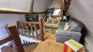 A cozy loft-style room with a wooden staircase, a grey sofa, a small table, and a colorful box on the floor. - La Chaumière aux Lamas (Goupil-Othon)