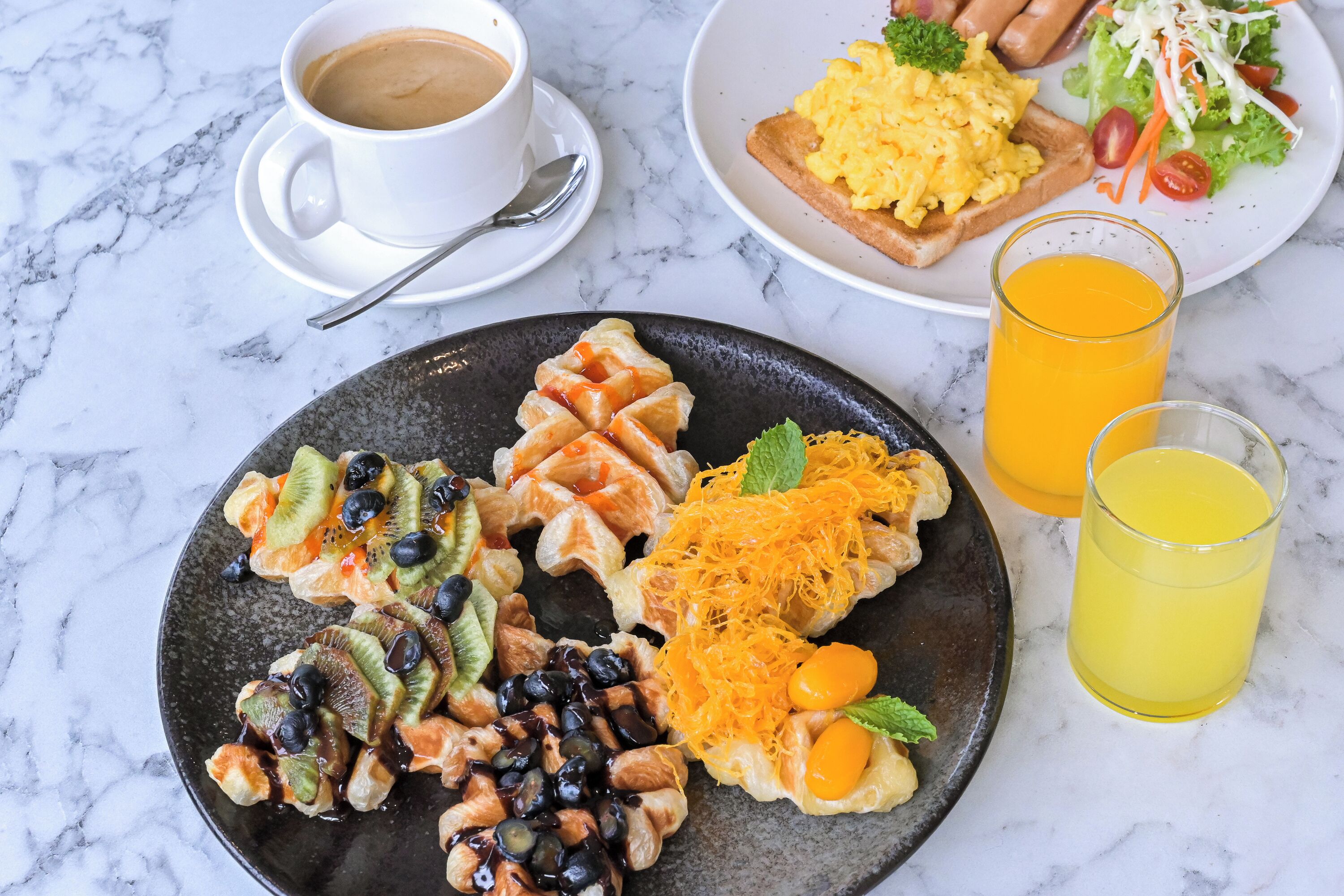 Un desayuno con un plato de pasteles variados, una taza de café, huevos revueltos, una ensalada y dos vasos de zumo de naranja sobre una mesa de mármol.