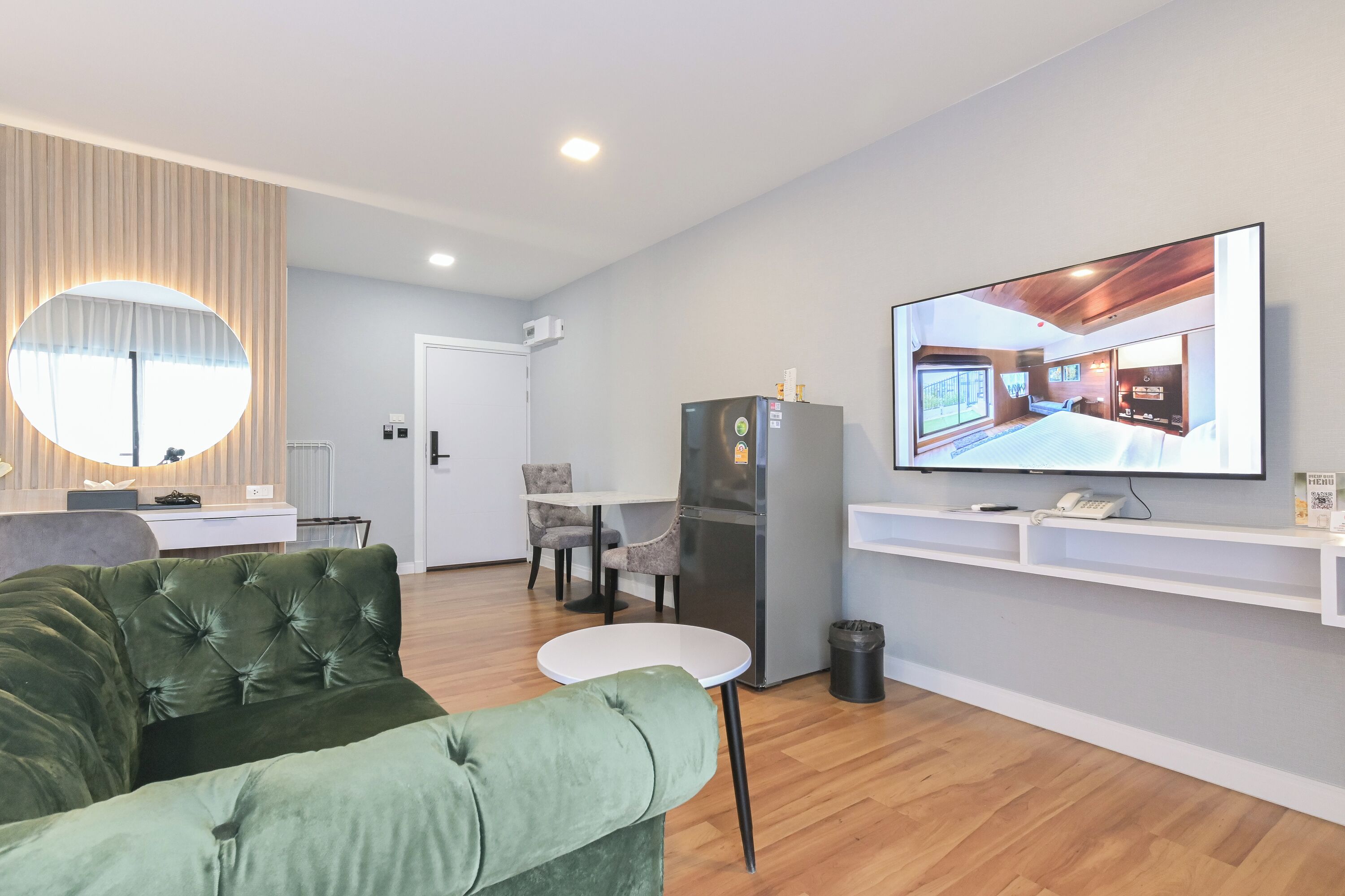 A modern living room with a green sofa, a round mirror, a flat-screen TV displaying a room, a refrigerator, and a small table.