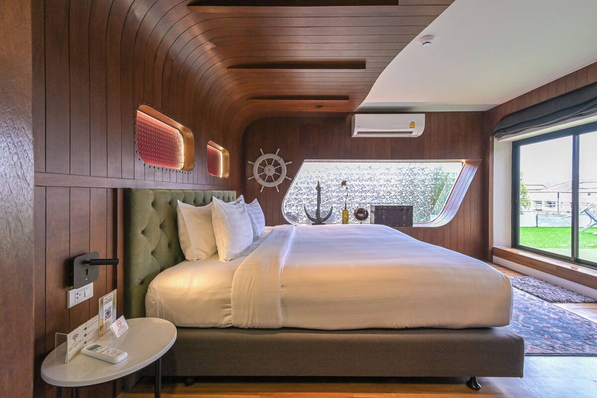 A modern bedroom with a large bed, a small bedside table, a wall-mounted clock, and a window with a view of greenery.
