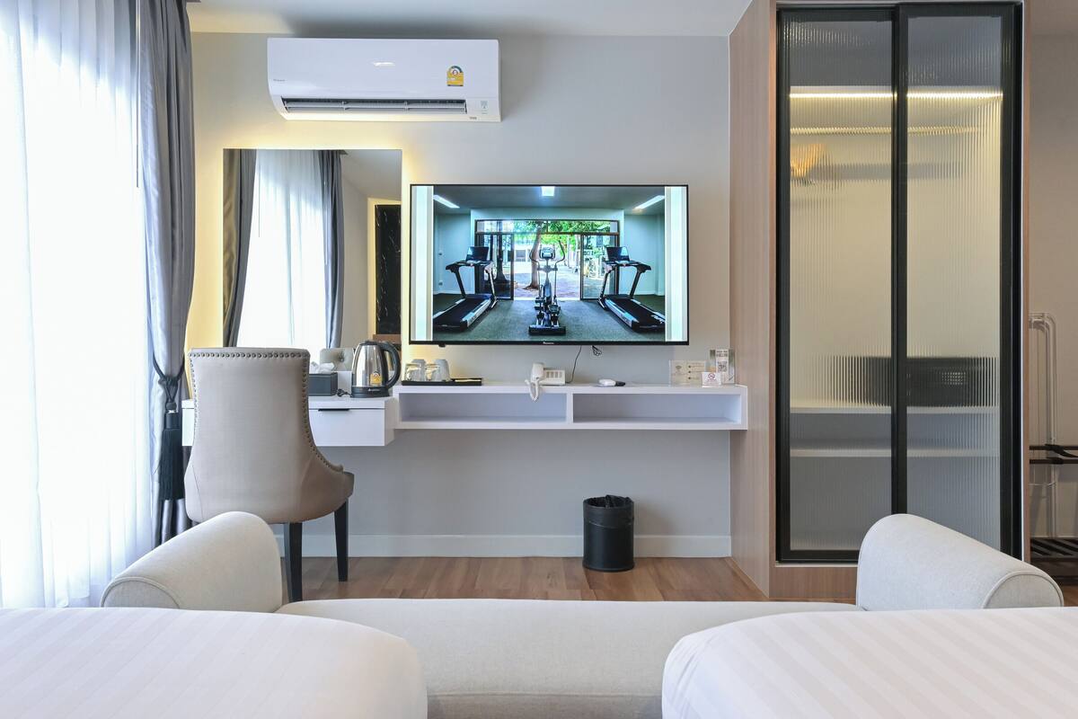 A hotel room with a TV displaying a gym, a desk with a chair, a trash bin, and a bed.