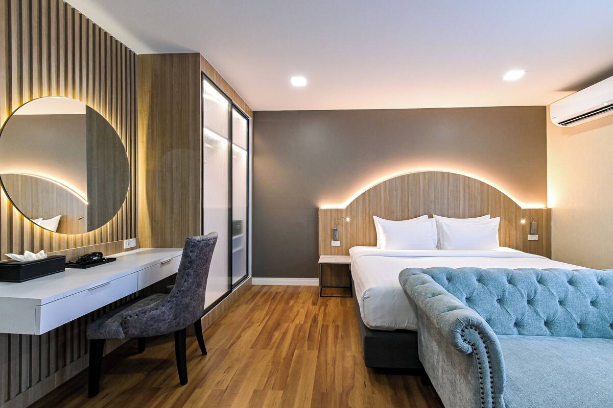 A modern hotel room with a wooden desk, a comfortable chair, a large bed with a tufted headboard, and a large mirror.