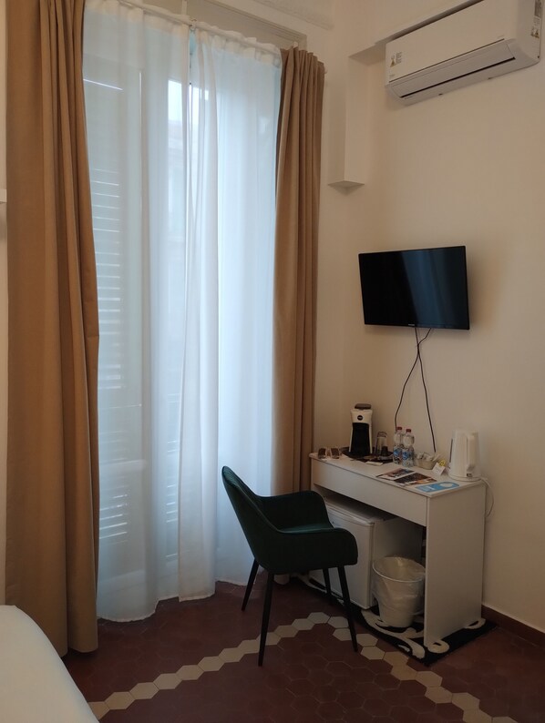 Habitación de hotel con cama, escritorio con silla, televisión, cafetera y cubo de basura.
