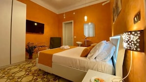 Habitación de hotel con cama, una mesita de noche con una bandeja de fruta, una planta en maceta y un espejo en la pared.