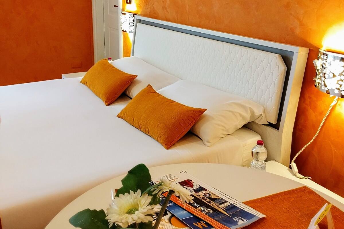 Een hotelkamer met een bed, een nachtkastje, een schilderij aan de muur en een tafeltje met een bloevas en tijdschriften.