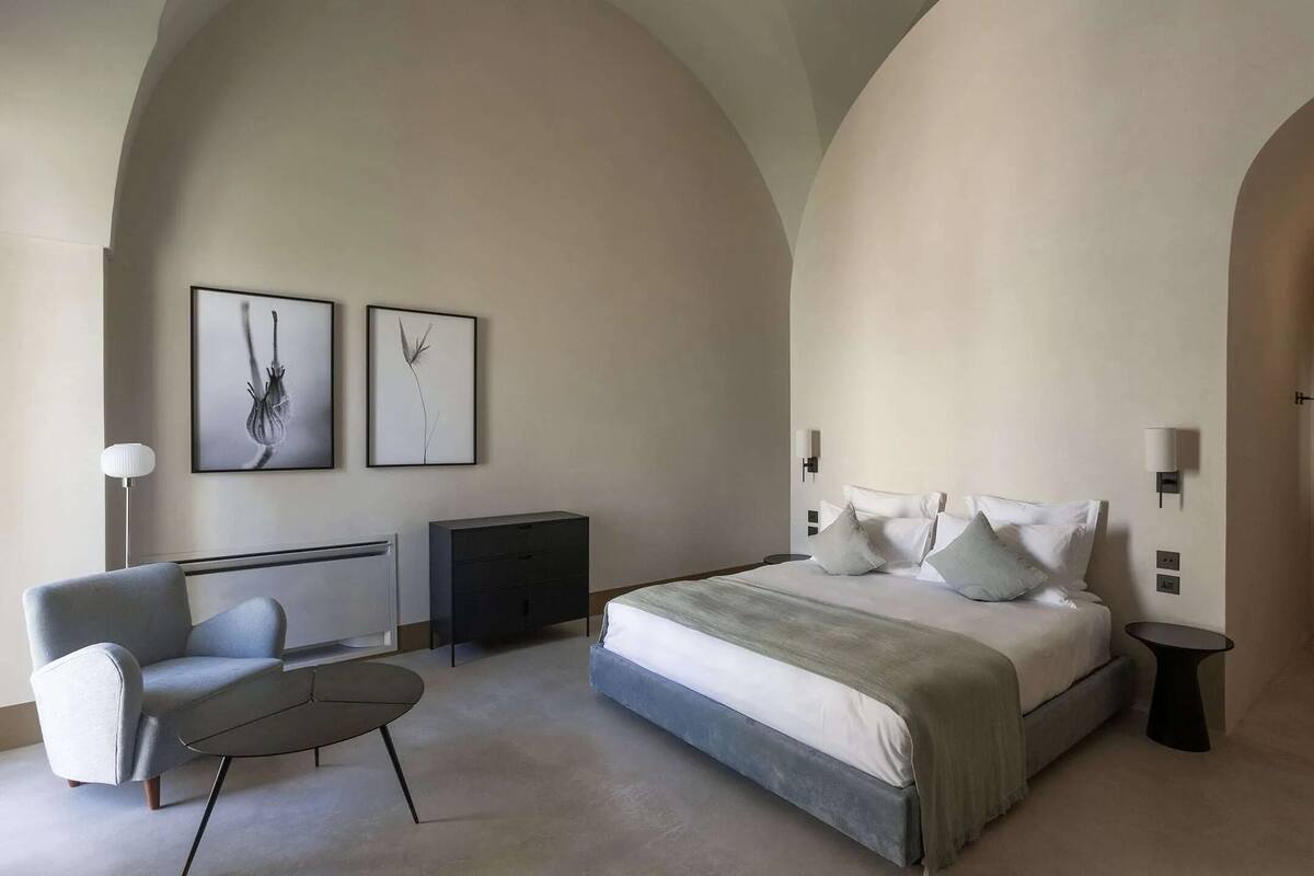 Un dormitorio moderno con una cama grande, una silla, una mesita y dos lámparas fijadas a la pared.