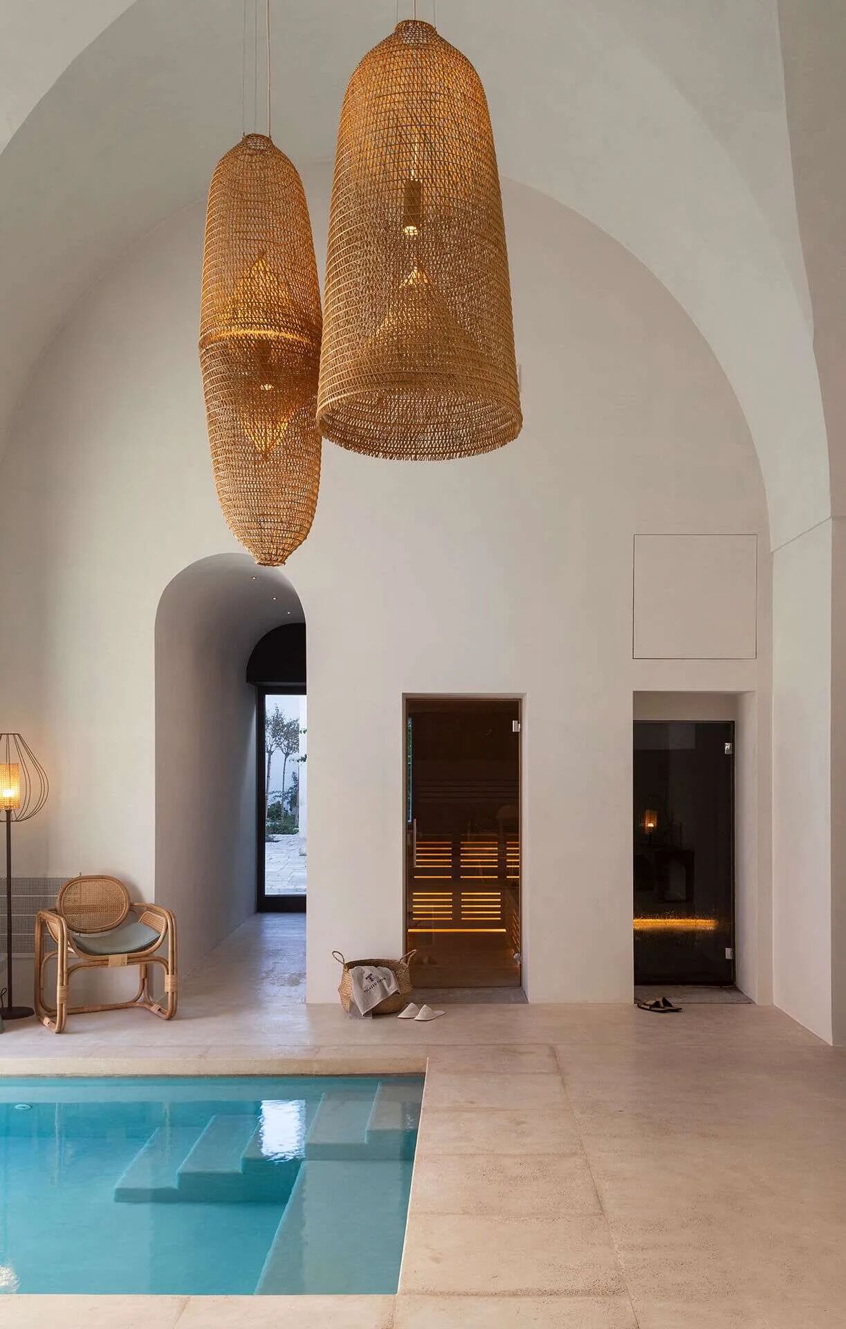 Un espacio interior amplio con piscina, lámparas colgantes de mimbre, una silla de mimbre y un acceso a otra habitación.