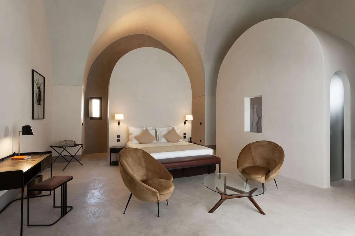 Una habitación de hotel moderna con una cama grande, dos sillones, un escritorio y una mesa de centro de vidrio.