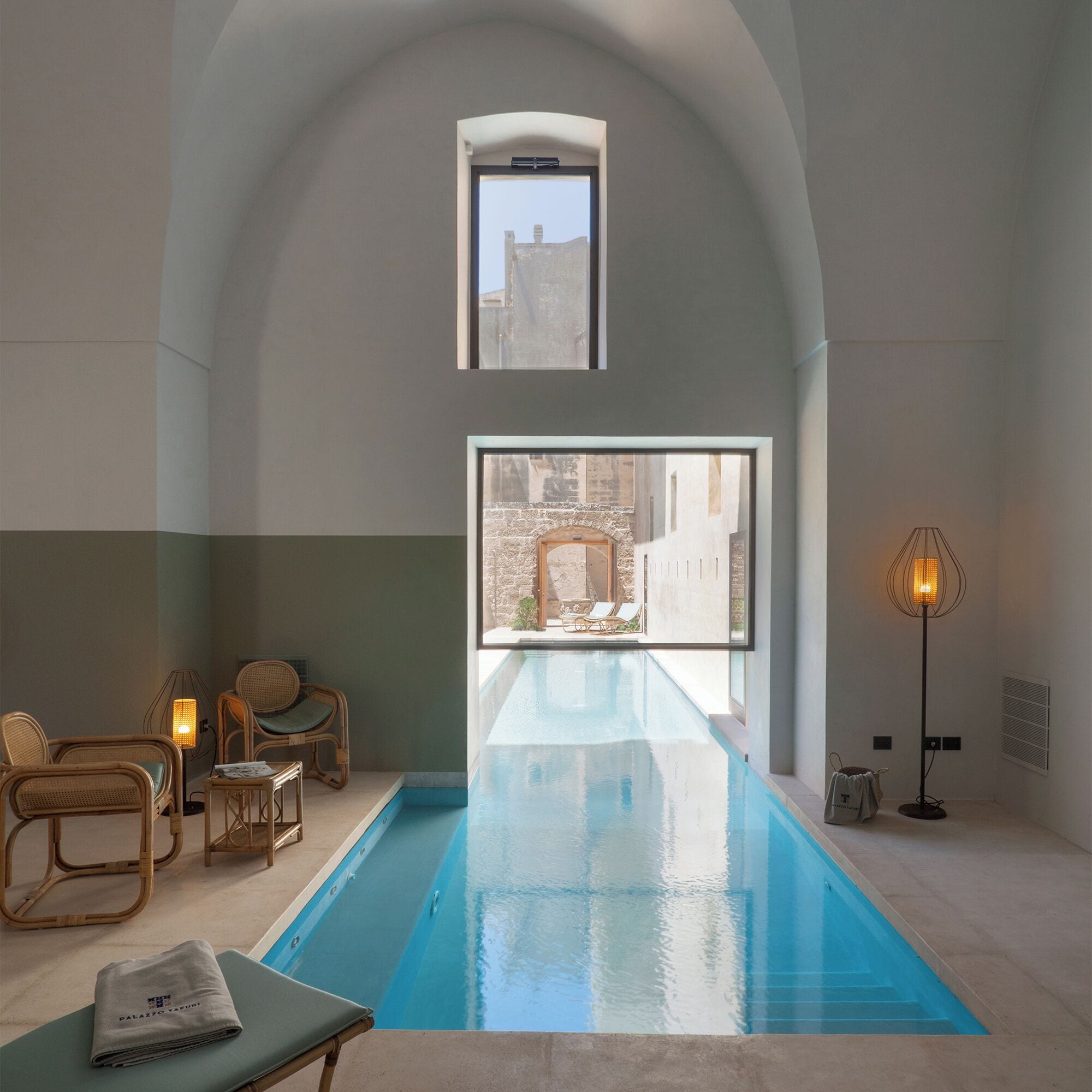 Un área de piscina interior moderna con vistas despejadas al exterior, mobiliario de mimbre y una lámpara de pie.