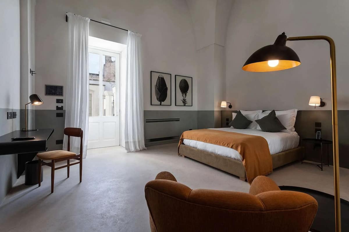 Una habitación de hotel moderna con cama, escritorio y silla, que cuenta con un ventanal grande y obras de arte en las paredes.