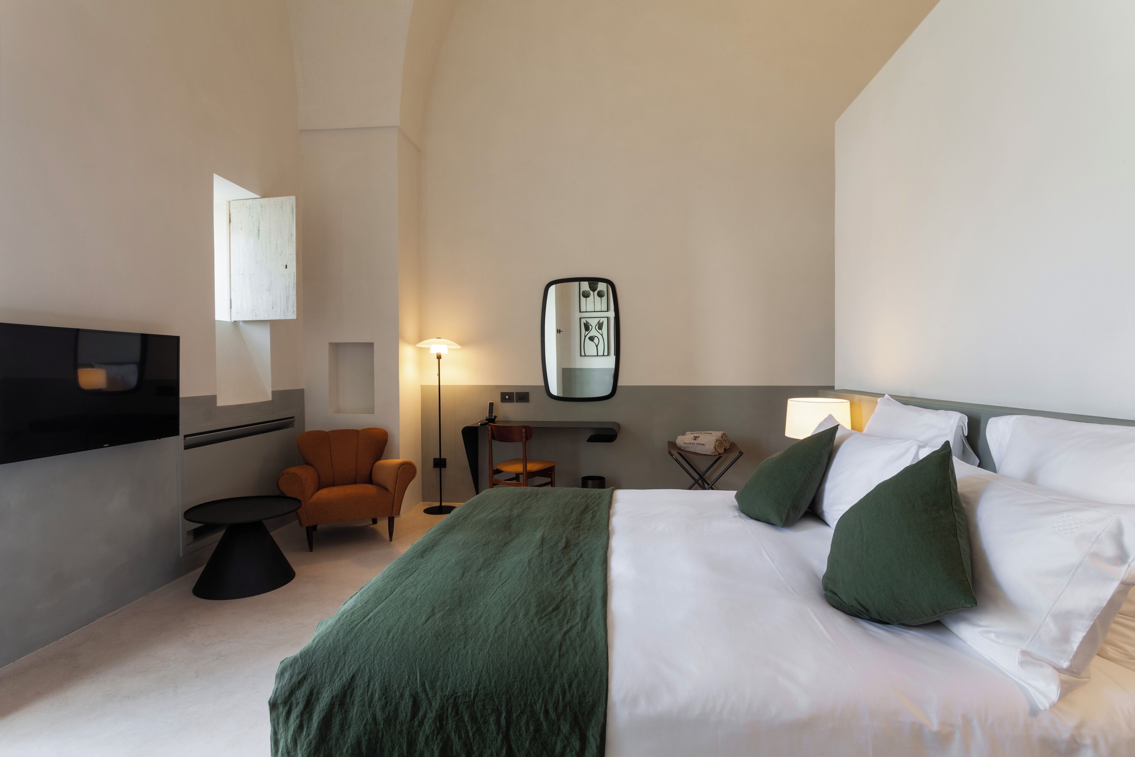 Habitación de hotel con cama, almohadas verdes, escritorio, silla, lámpara y espejo en la pared.