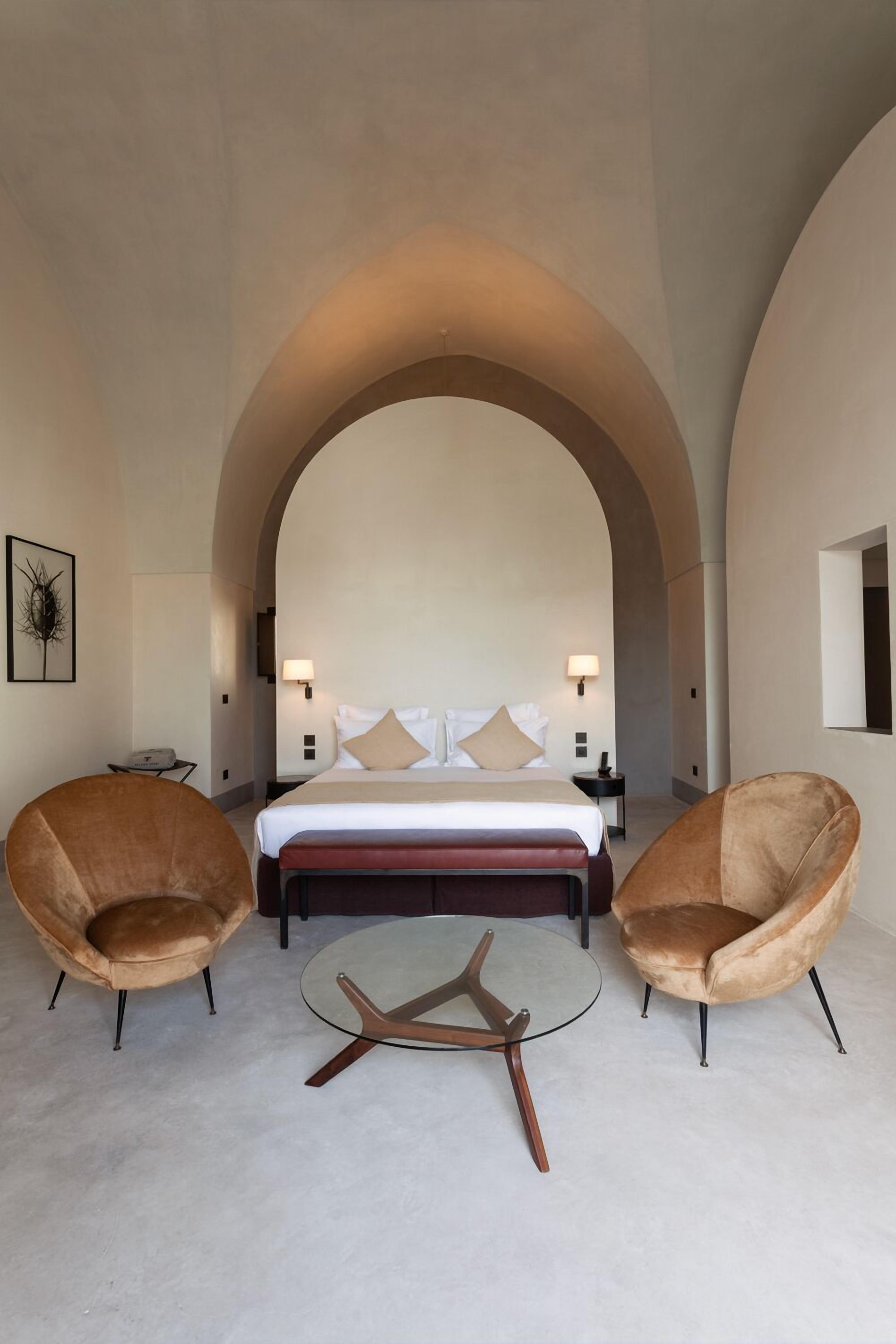 A hotel room with a large bed, two armchairs, a small round table, and a large arched doorway.