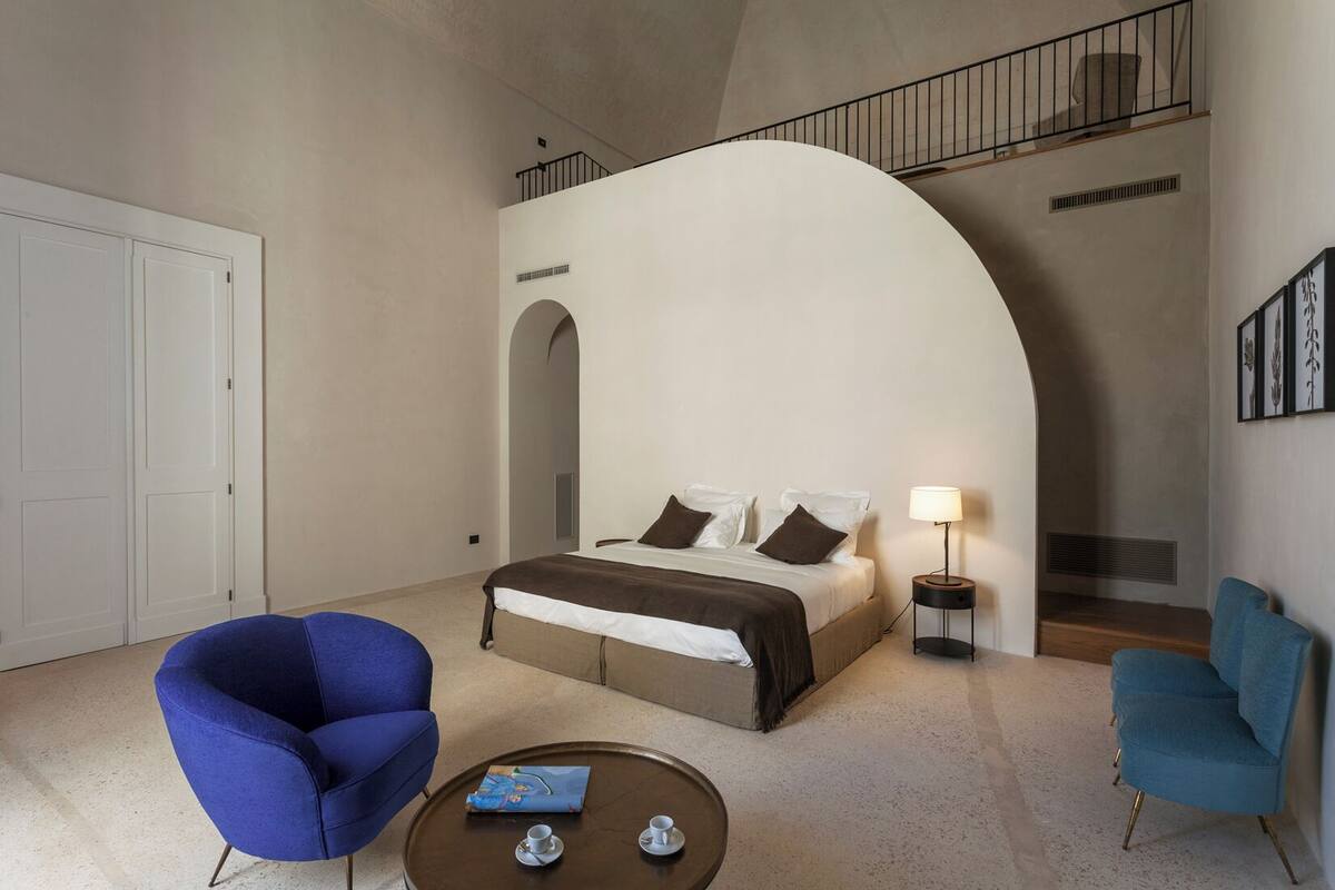 Un dormitorio moderno con una cama grande, dos sillones azules, una mesita de centro con tazas y una escalera con barandilla.