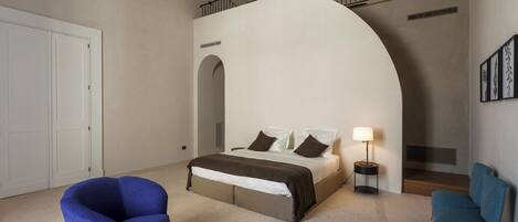 A modern bedroom with a large bed, two blue armchairs, a coffee table with cups, and a staircase with a railing.