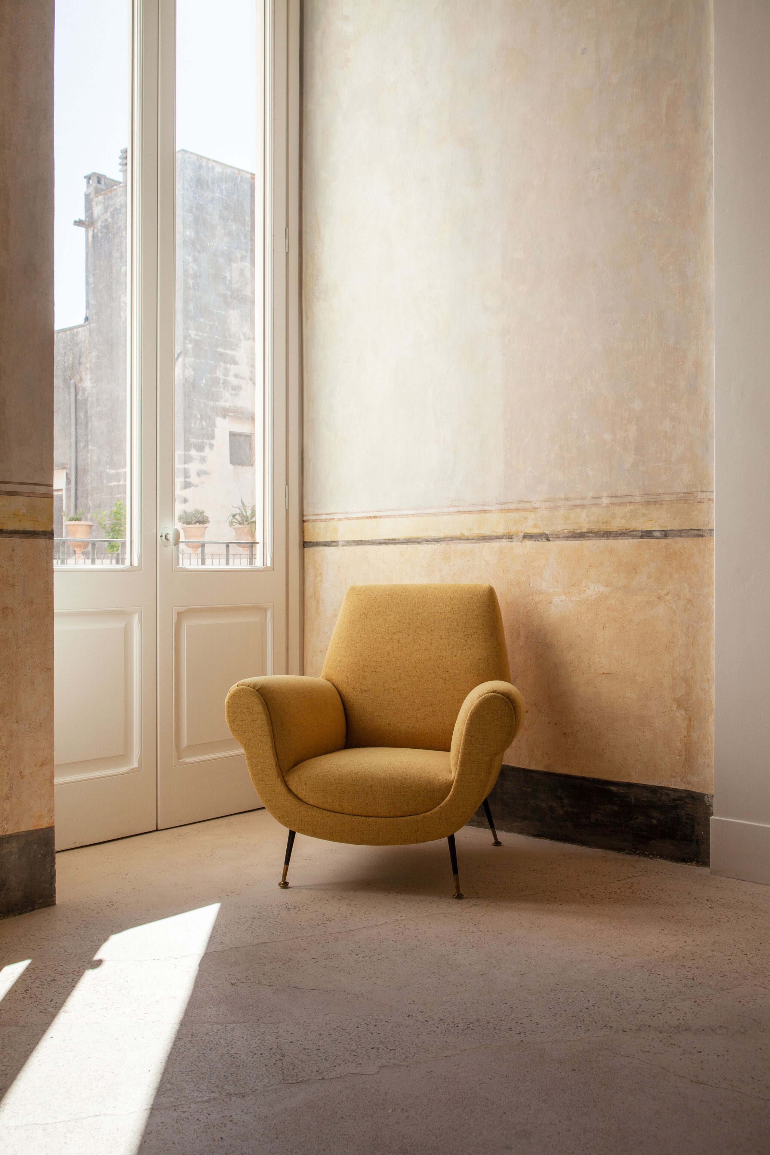 A mustard-colored armchair in a room with a large window, a textured wall, and a door.
