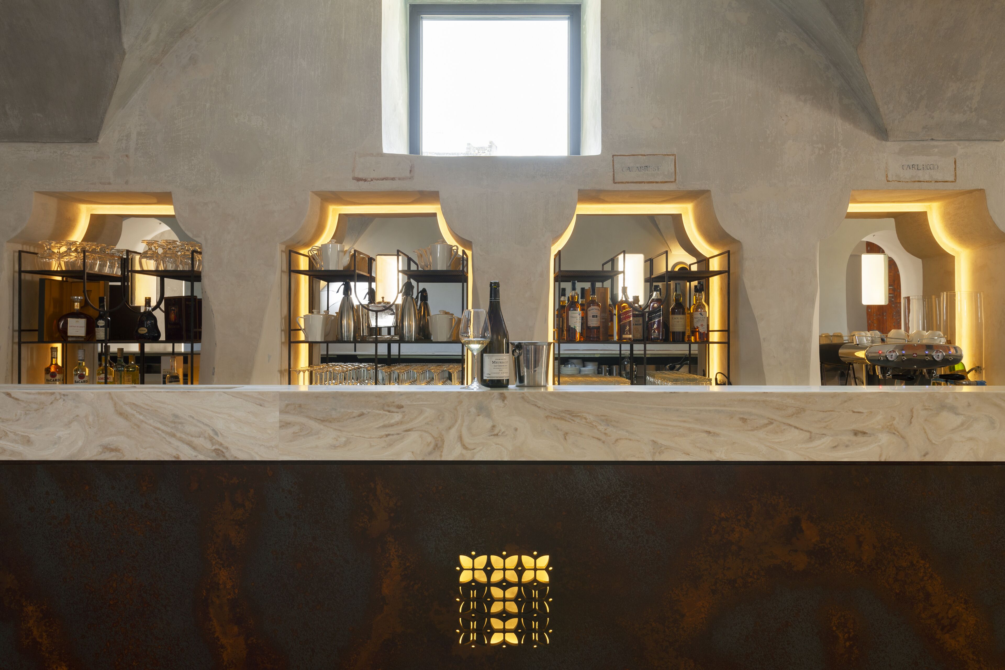 A bar area with shelves of bottles and glasses, a marble countertop, and a window above.