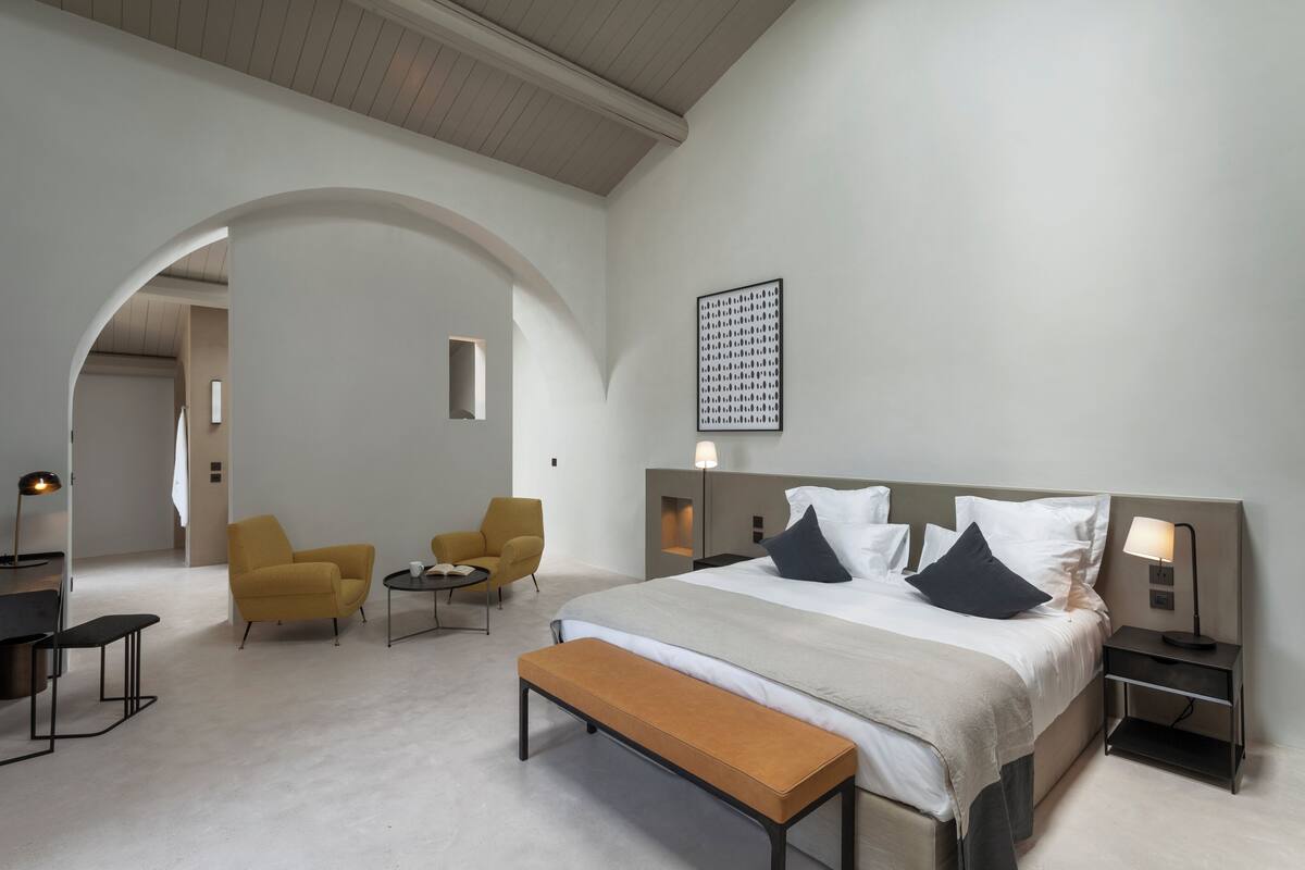 Una habitación de hotel moderna con una cama grande, un banco, dos sillones, una mesita y una obra de arte colgada en la pared.