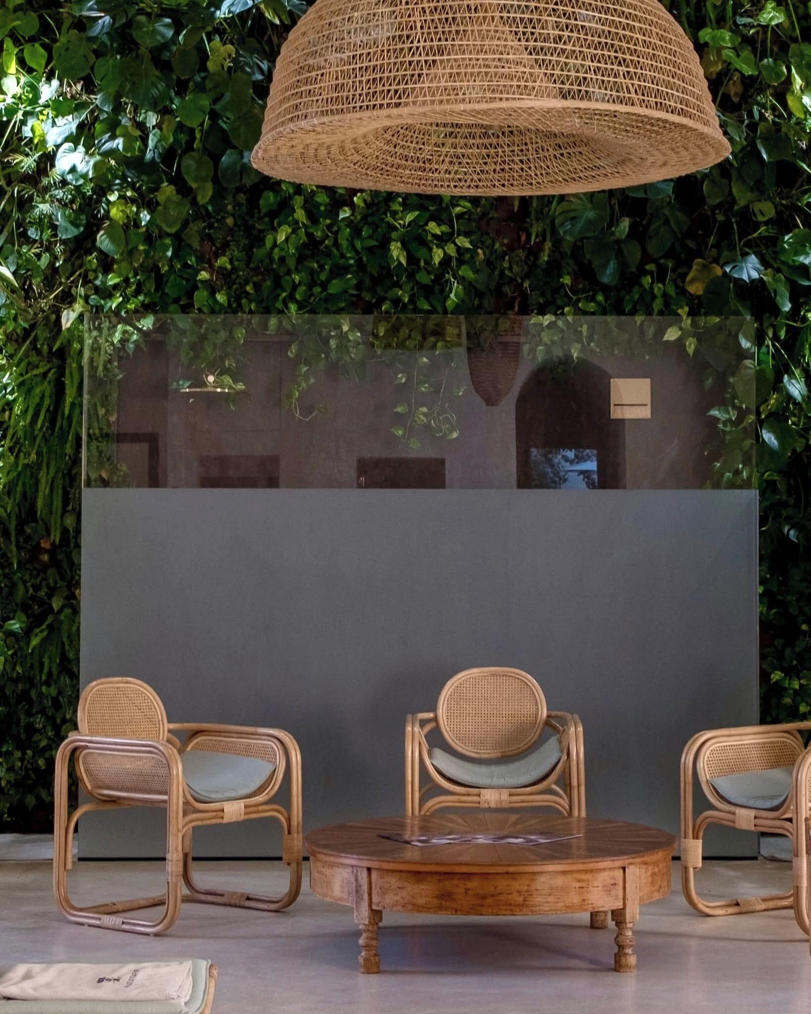 A room with a round wooden table and two wicker chairs, a woven ceiling light, and a glass wall with greenery behind it.