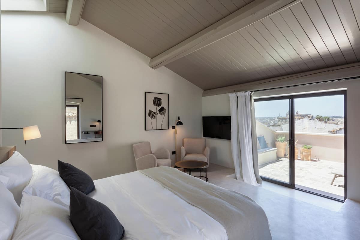 Habitación de hotel con una cama grande, una silla, una mesita, un televisor y un balcón con plantas en macetas.