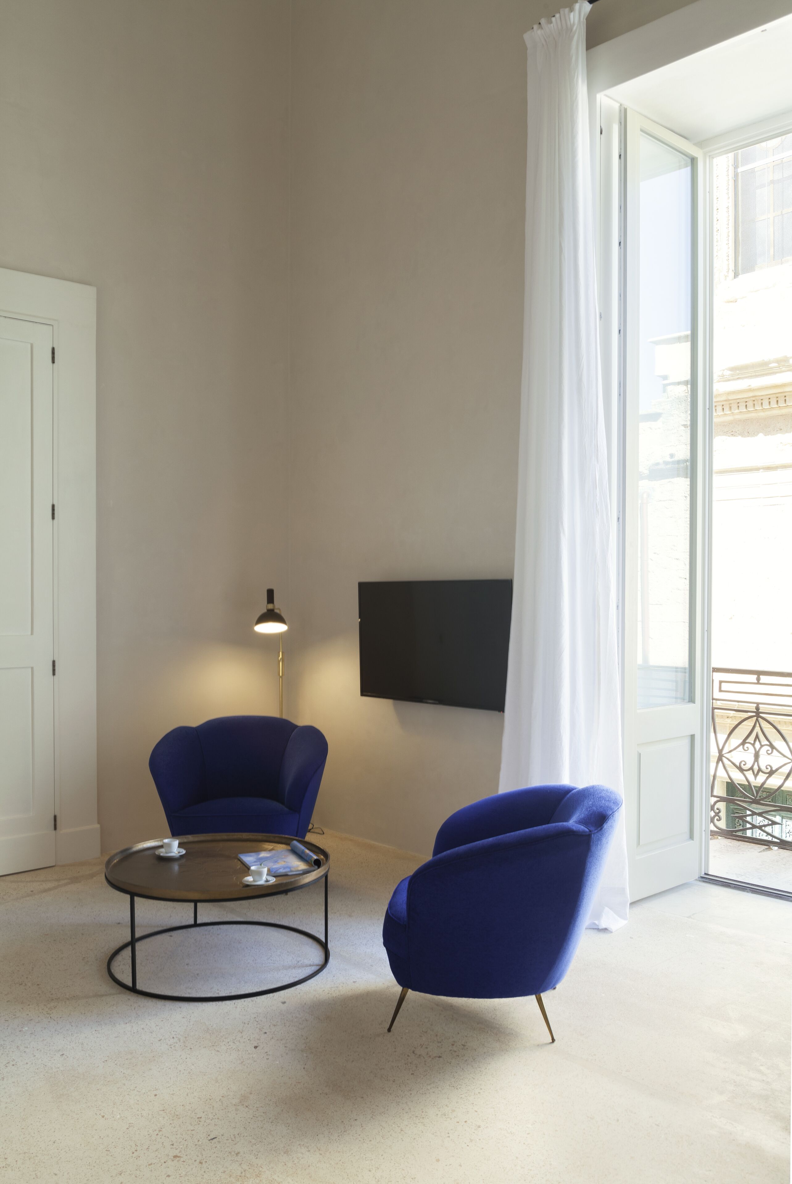 Una habitación moderna con dos sillones azules, una mesita redonda pequeña, una lámpara de pie y un televisor montado en la pared.