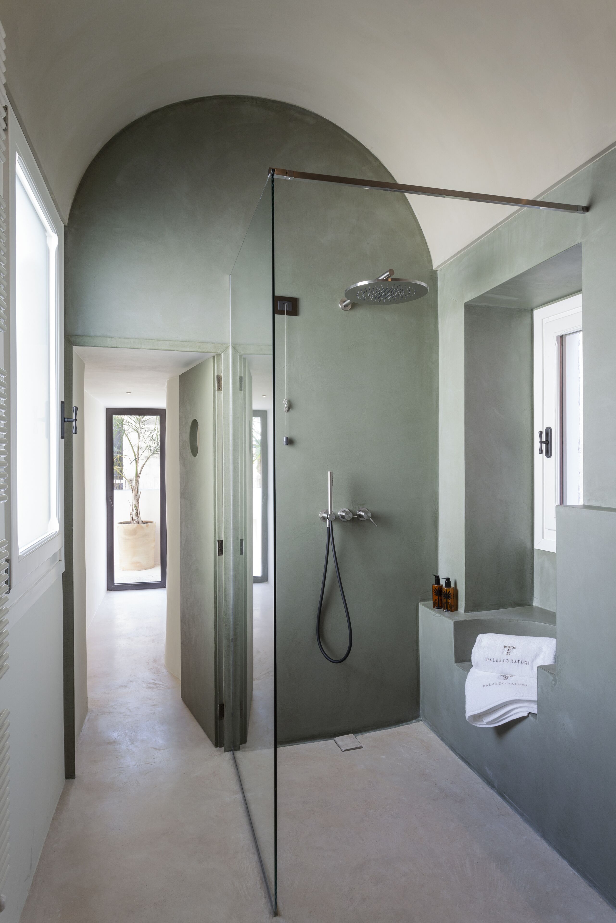 Un baño moderno con ducha de vidrio, un espejo grande y vista a un árbol a través de una puerta abierta.
