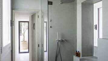 Un baño moderno con ducha de vidrio, un espejo grande y vista a un árbol a través de una puerta abierta.