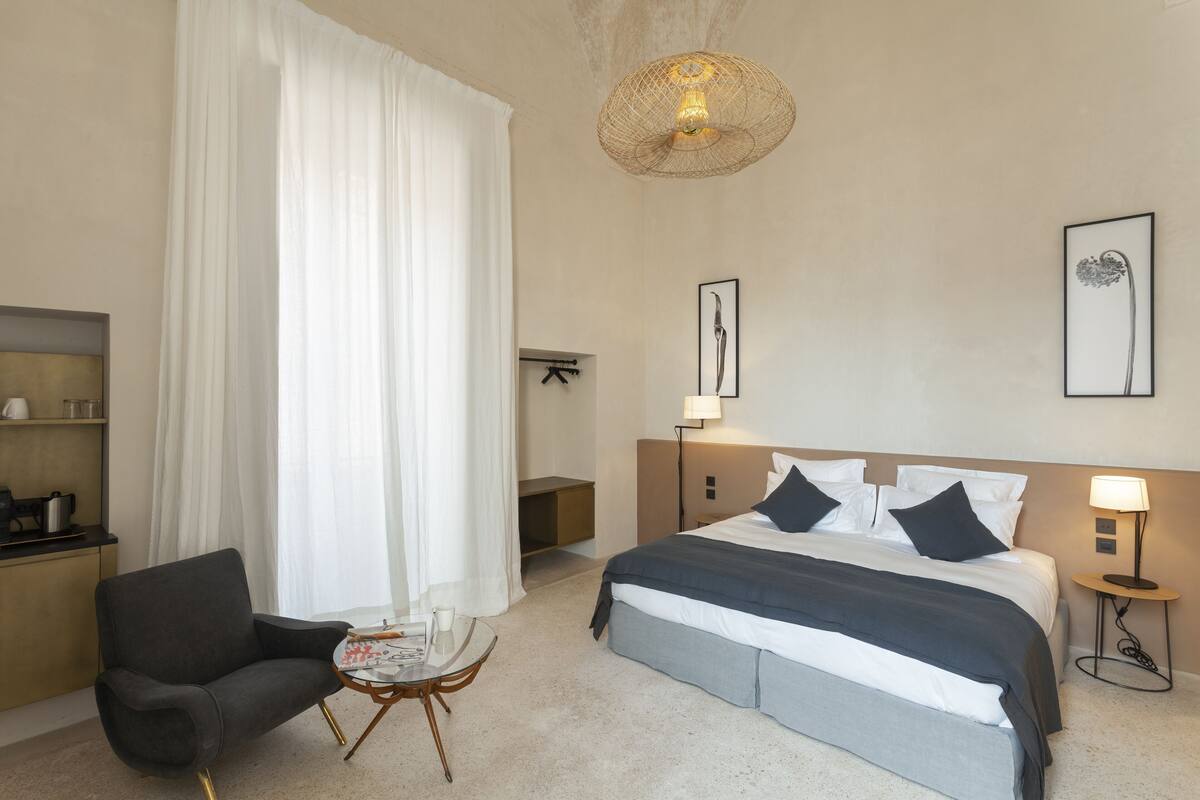 Una habitación de hotel con cama, una silla, una mesita, una lámpara y obras de arte en las paredes.