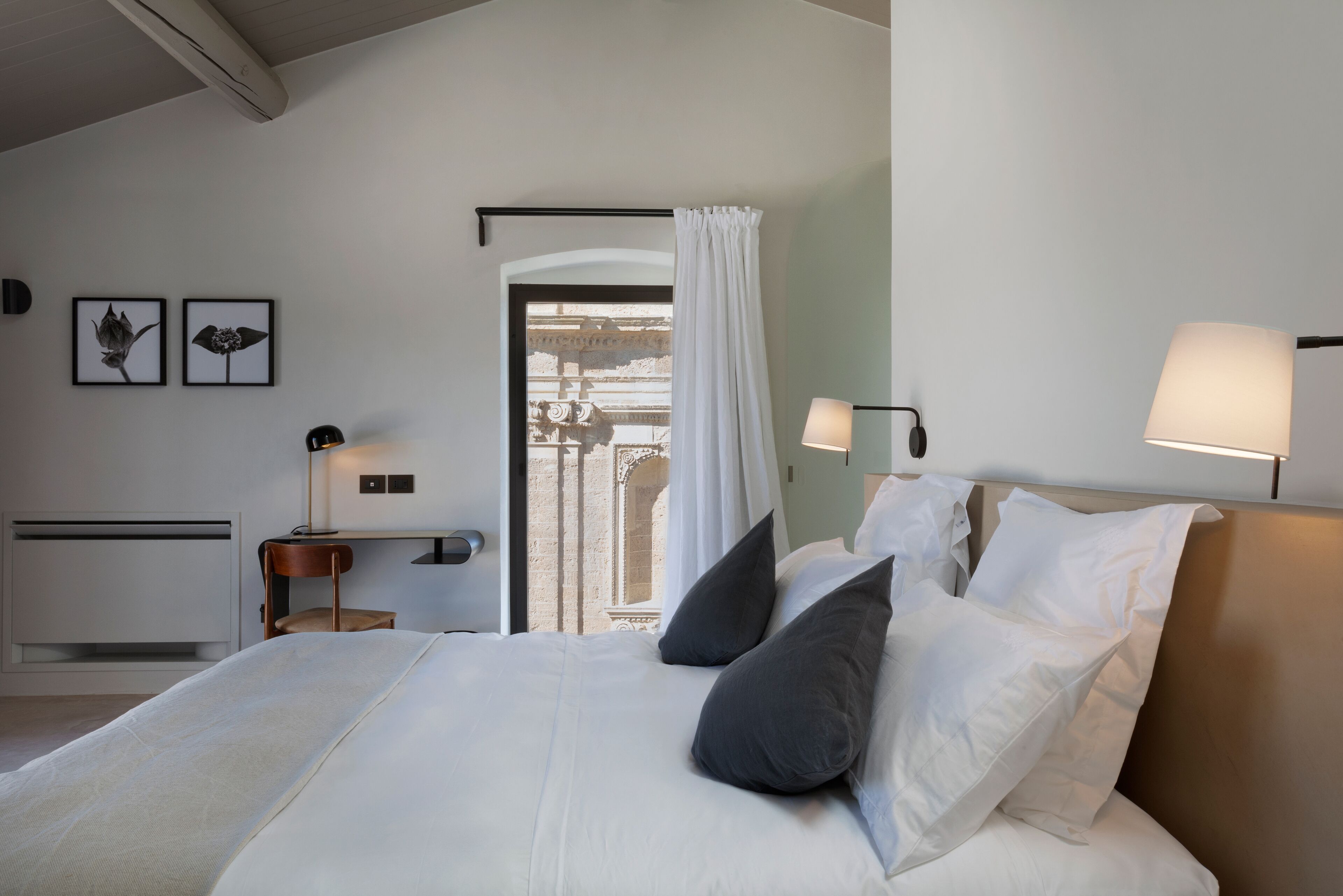A modern hotel room with a large bed, two bedside lamps, and a view of a courtyard through a window with white curtains.