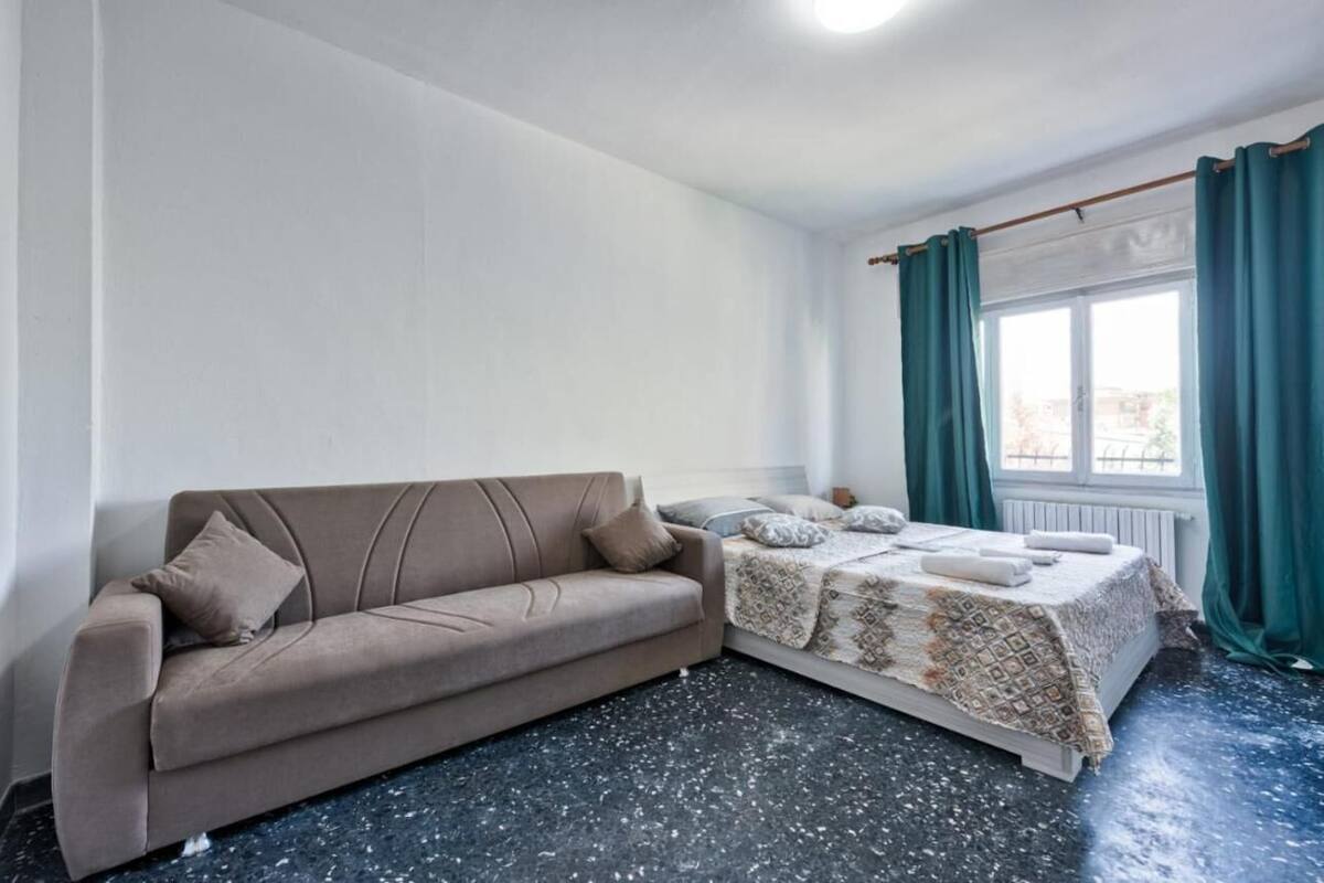 A room with a sofa, a bed with a patterned bedspread, a window with curtains, and a radiator.