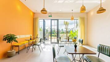 Uma sala de jantar luminosa e moderna, com uma grande janela, várias mesas e cadeiras, e luminárias pendentes.