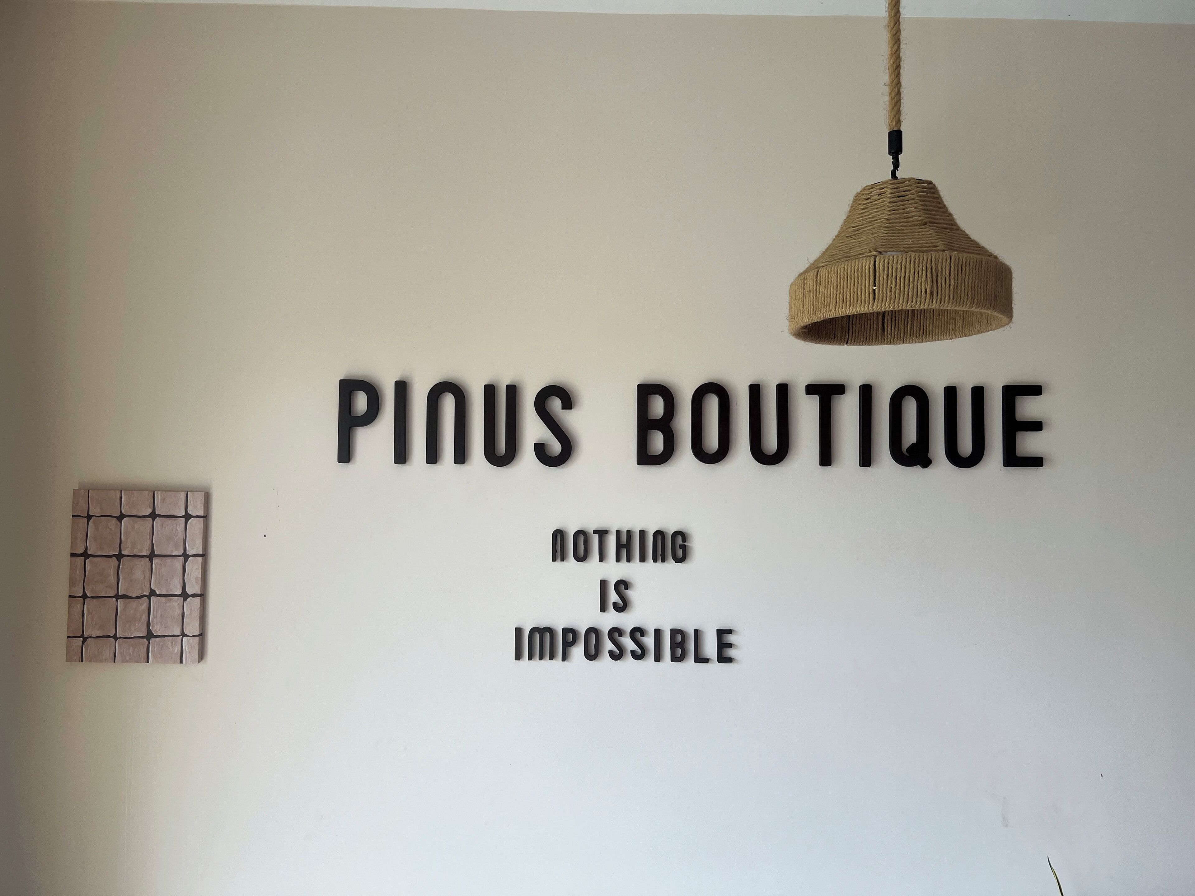 Seinässä on mustilla kirjaimilla kirjoitettu teksti: ”PINUS BOUTIQUE” ja ”MIKÄÄN EI OLE MAHDOTONTA”. Lisäksi seinässä on punottu riippuvalaisin ja pieni, neliönmuotoinen koriste.