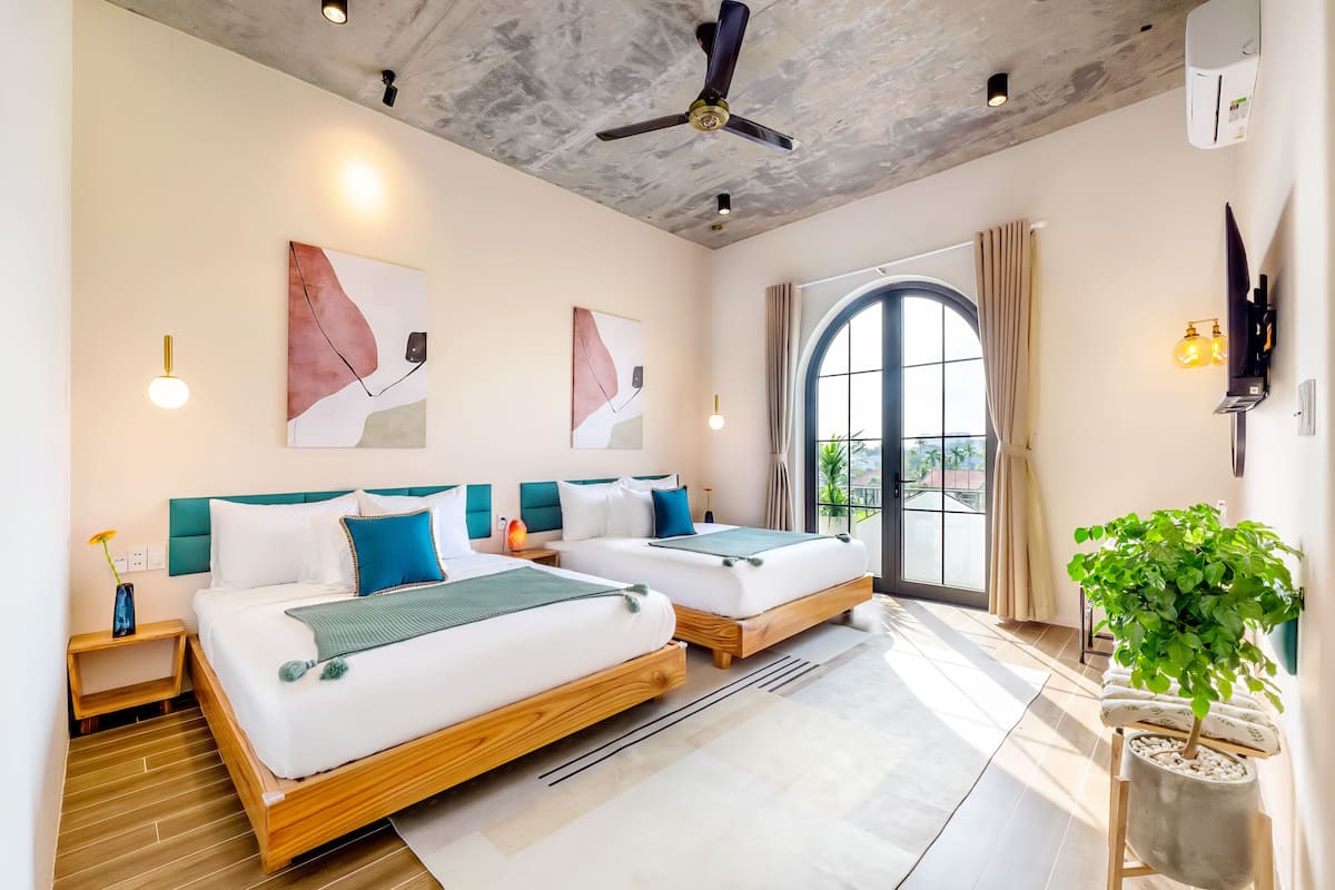 A modern hotel room with two beds, a ceiling fan, artwork on the walls, a potted plant, and a large window with curtains.