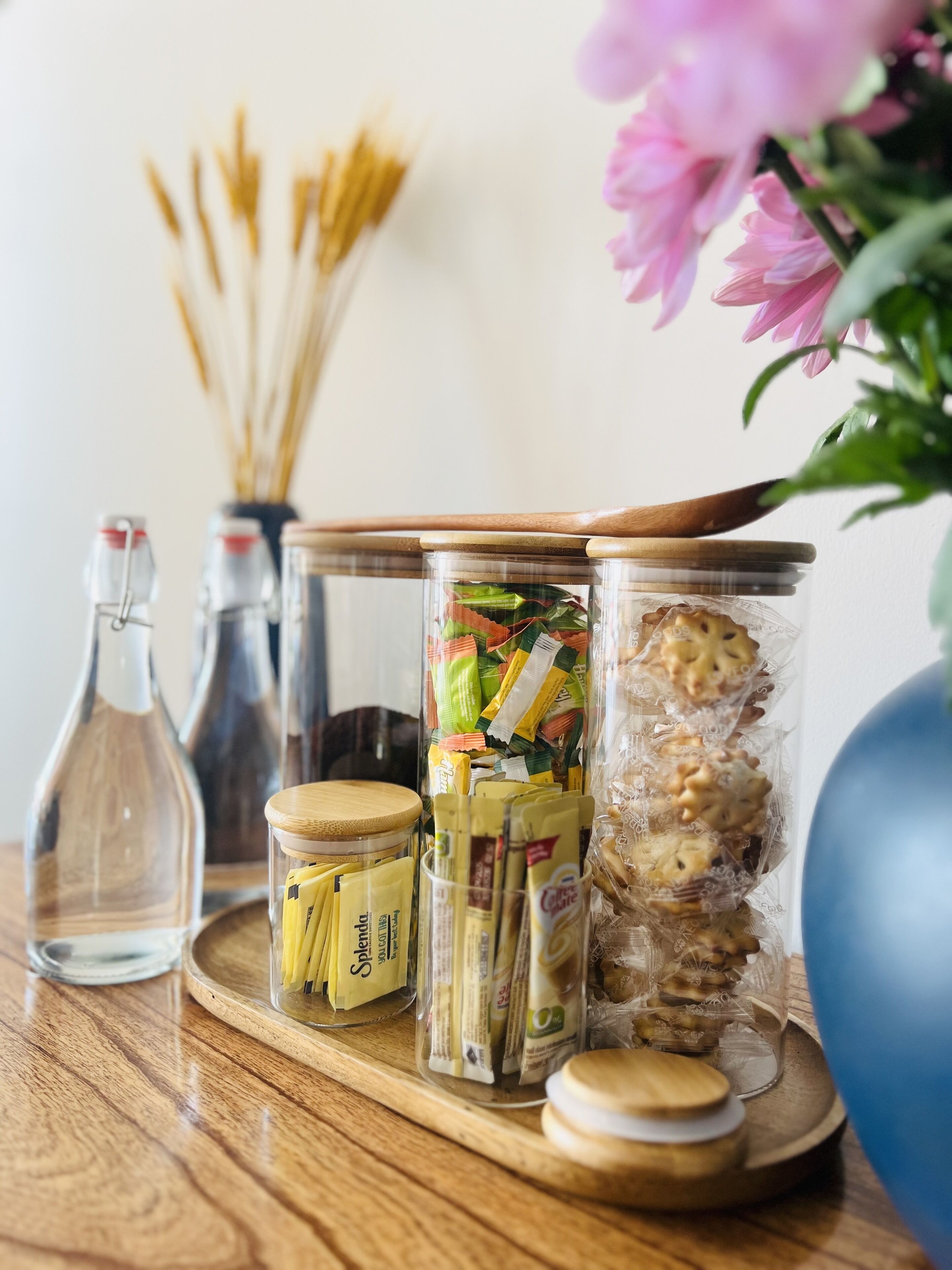 En træbakke med glasbeholdere med snacks, en flaske og en vase med blomster.