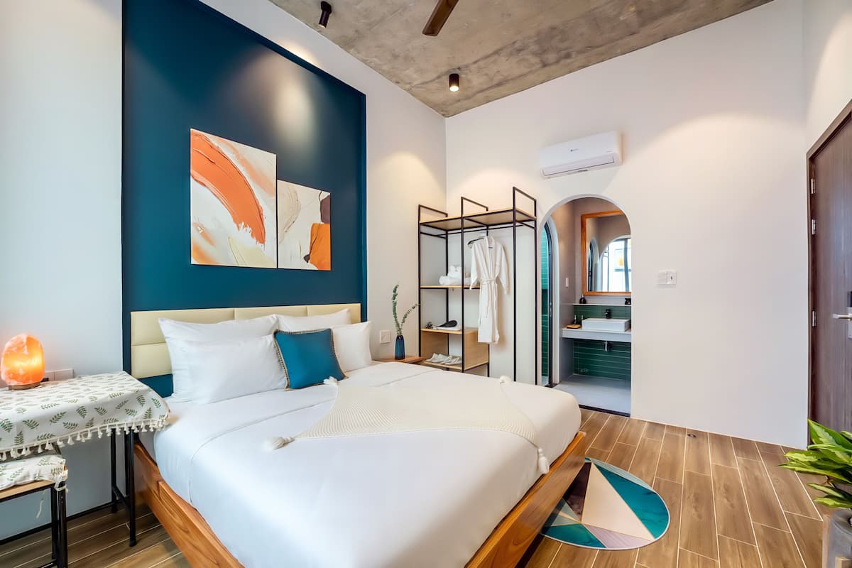 A modern bedroom with a large bed, a bedside table with a lamp, and a wall-mounted shelf with clothes.
