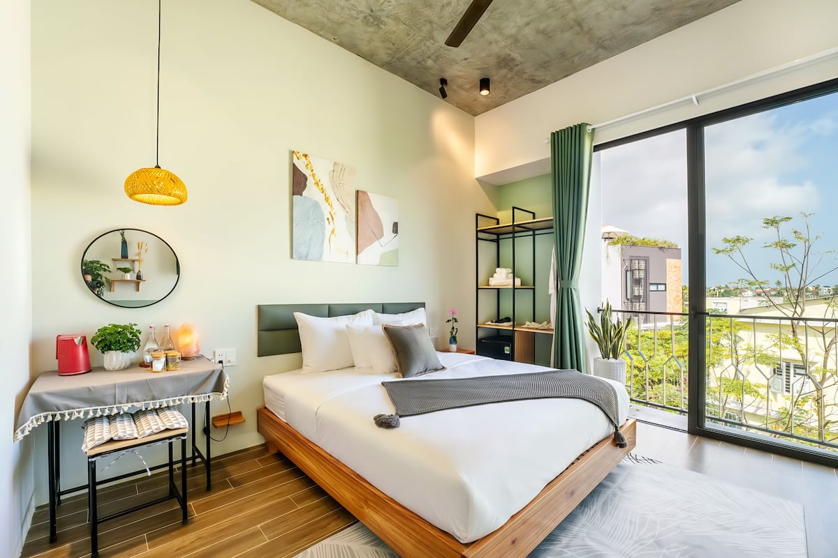 A modern bedroom with a large bed, a bedside table with a lamp, a mirror, artwork on the wall, and a view of the city through a large window.