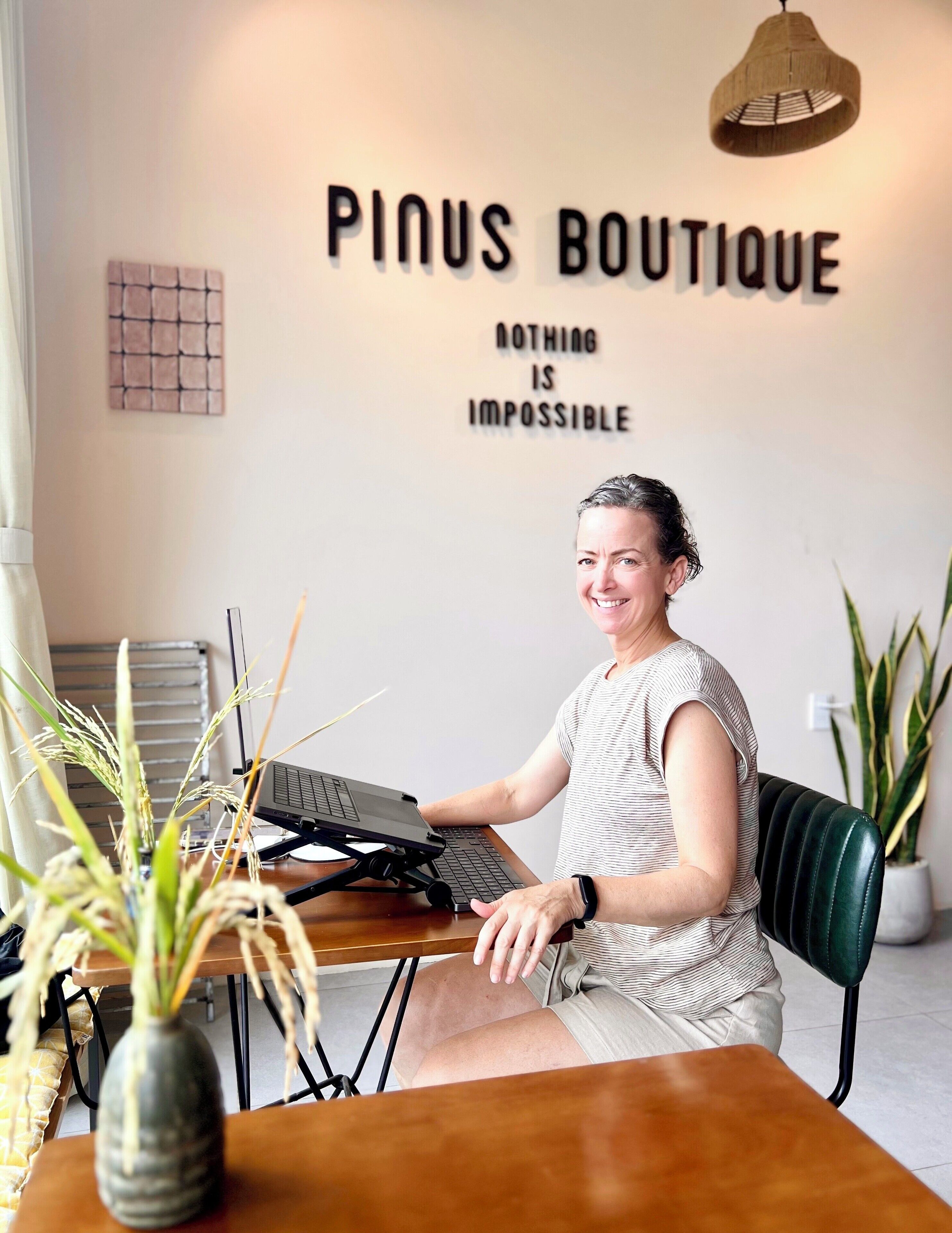 Una persona sentada en un escritorio con una computadora portátil en una habitación con un letrero que dice "PINUS BOUTIQUE" y "NOTHING IS IMPOSSIBLE".