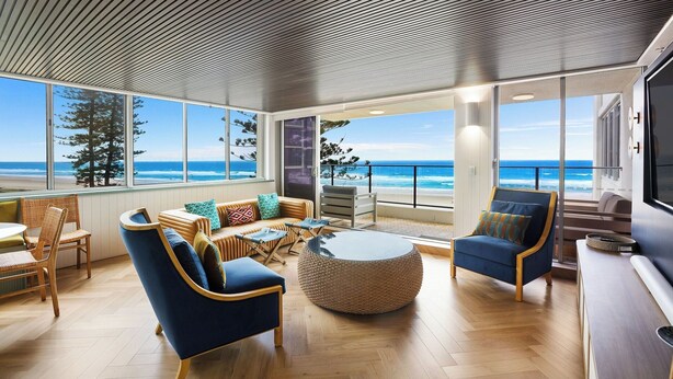 A spacious living area with a view of the beach, featuring a round coffee table, blue armchairs, and a wicker sofa.