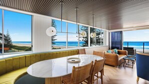 A modern dining area with a large oval table, wicker chairs, and a view of the beach and ocean through large windows.