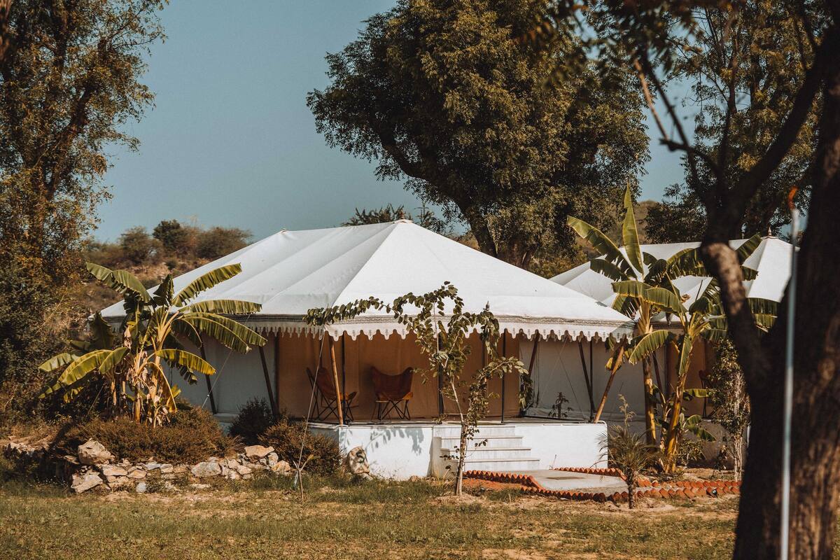 A white tent-like structure with a flat roof, surrounded by trees and a small garden area with a stone wall and steps leading up to the entrance.