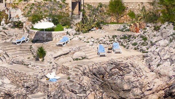 Ett hotell vid kusten med en terrass av sten, solstolar och en fri utsikt över havet.