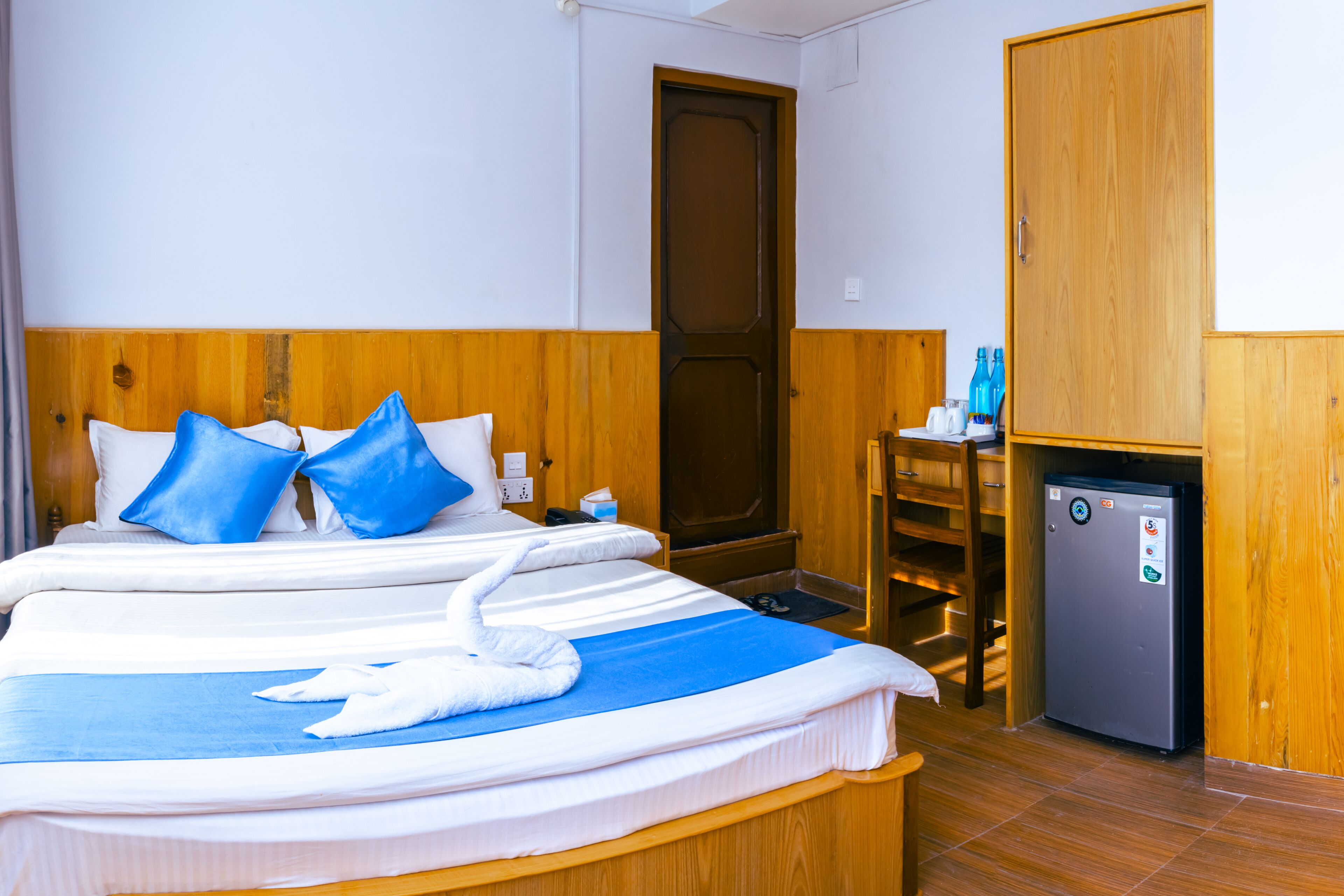 A hotel room with a large bed, wooden headboard, a small refrigerator, and a wooden wardrobe.