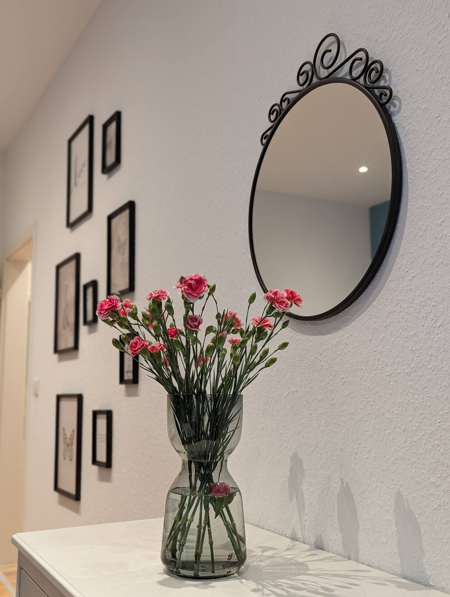 Et badeværelse med en hvid kommode, en vase med lyserøde blomster og et rundt spejl med dekorativ ramme.