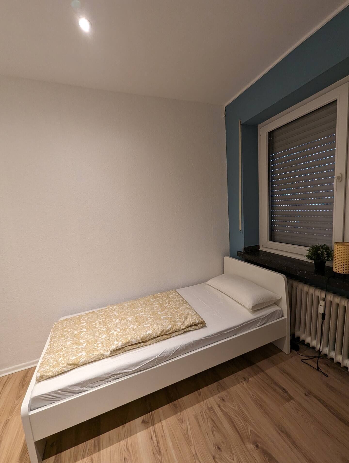 Une petite chambre avec un lit simple, un plancher en bois, une fenêtre avec des stores et un radiateur.