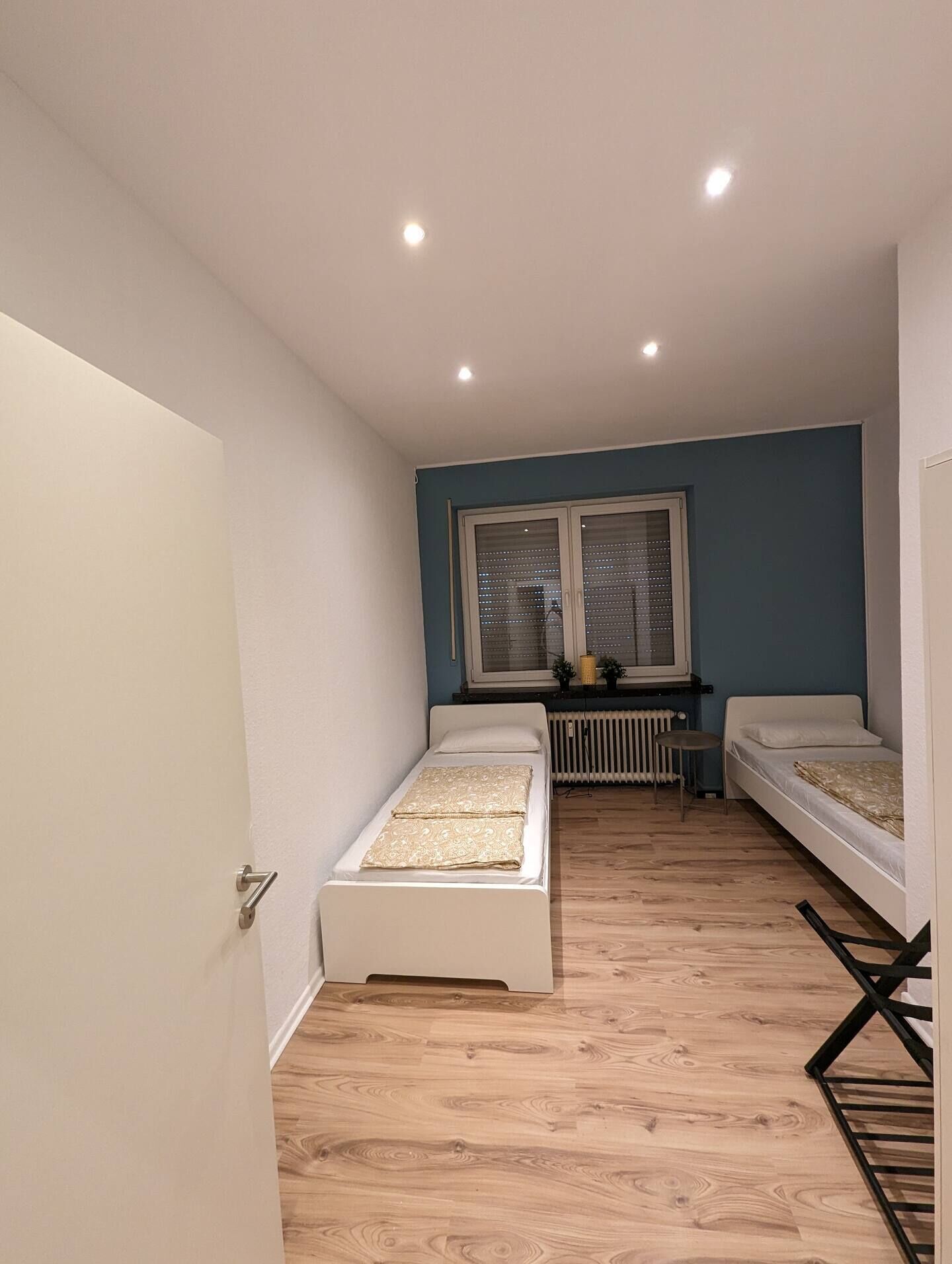Une petite pièce bien éclairée, avec deux lits simples, une fenêtre avec des stores, un radiateur et un plancher en bois.