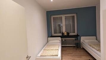 Une petite pièce bien éclairée, avec deux lits simples, une fenêtre avec des stores, un radiateur et un plancher en bois.