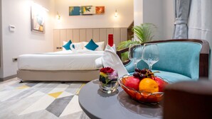 Une chambre d’hôtel avec un lit, un fauteuil bleu, une petite table avec un verre de vin, un vase rempli de fleurs et un panier de fruits.