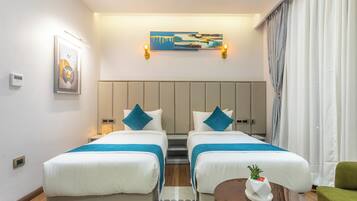 A hotel room with two beds, a small table, a green chair, and a painting on the wall.