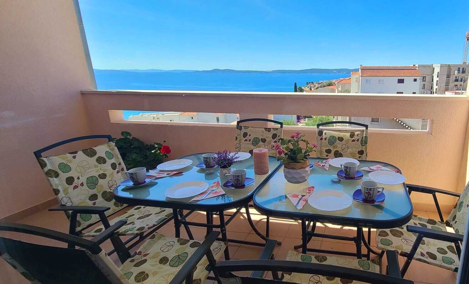 A balcony with a table set for two, chairs with leaf-patterned upholstery, and a view of the sea and buildings in the background.