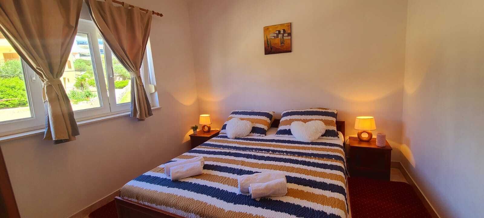 Hotellihuone, jossa on sänky, kaksi yöpöytälamppua, ikkuna verhoilla ja taulu seinällä.